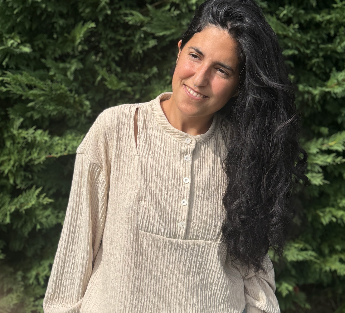 Mujer con cabello rizado y oscuro sonriendo, usando blusa beige, de fondo árboles verdes.