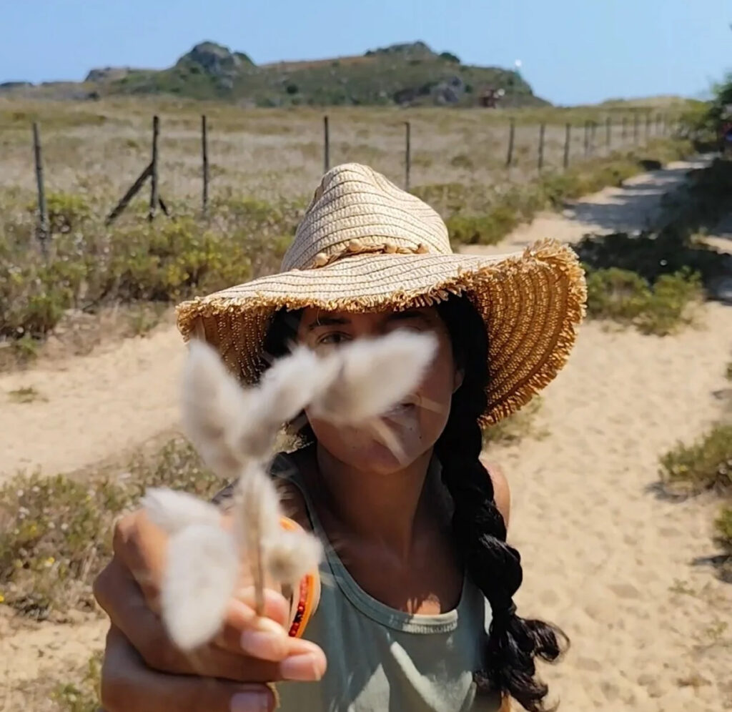 Mujer con sombrero de paja sostiene una rama con algodón en un paisaje árido con camino y colinas al fondo.